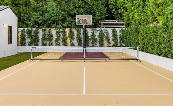 Modern backyard sports court with basketball hoop and net, surrounded by lush greenery and a small white building.