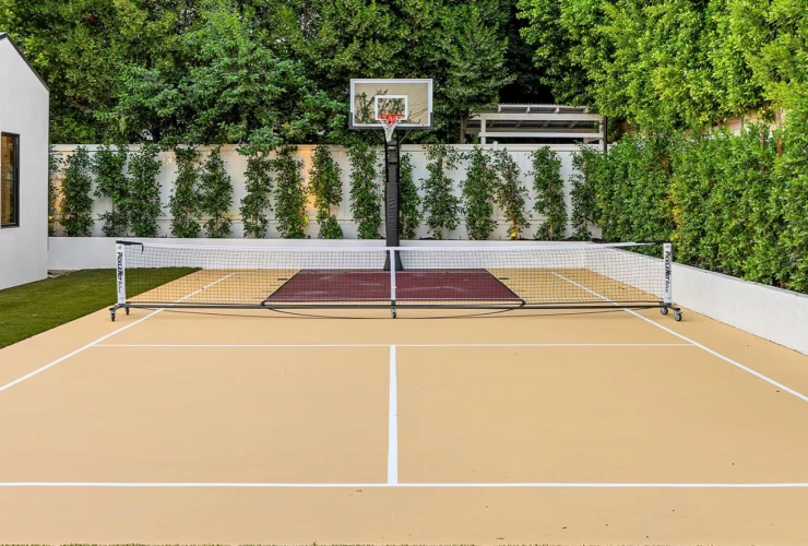 Modern backyard sports court with basketball hoop and net, surrounded by lush greenery and a small white building.