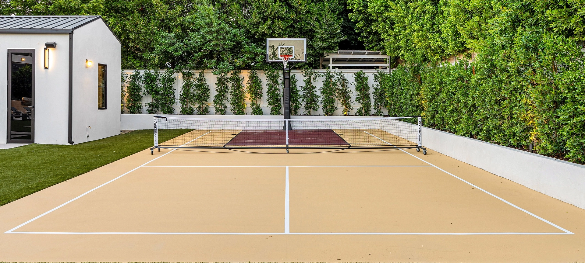 Modern backyard sports court with basketball hoop and net, surrounded by lush greenery and a small white building.