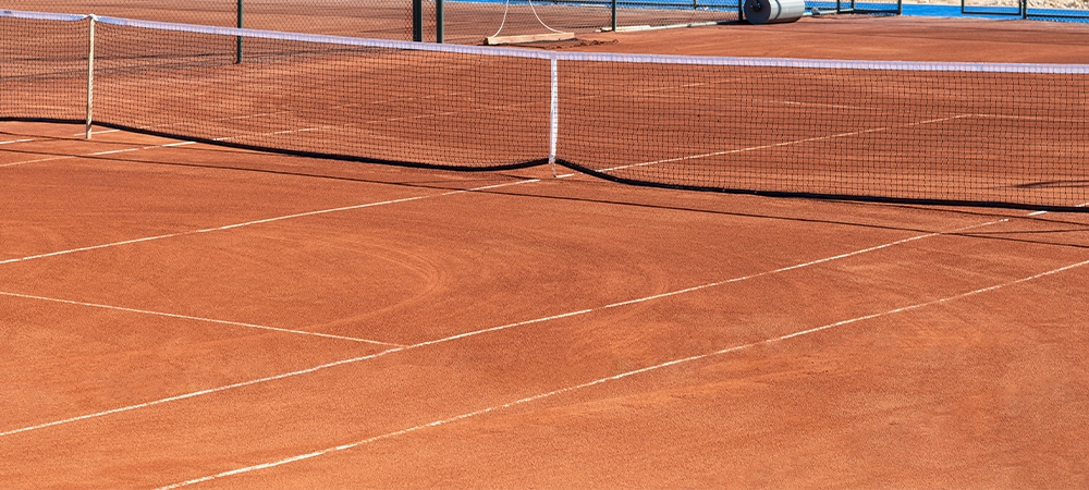 baseline and net of an empty clay tennis court on a sunny day