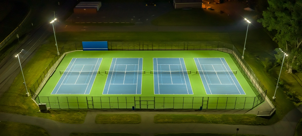 Evening aerial photo of outdoor blue tennis courts with pickleball lines and green out of bounds area with lights turned on.