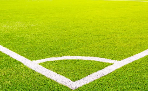 Corner of a green artificial turf soccer field with white lines. Professional football stadium background under bright sunlight. Concept of sports, competition, and outdoor activity.
