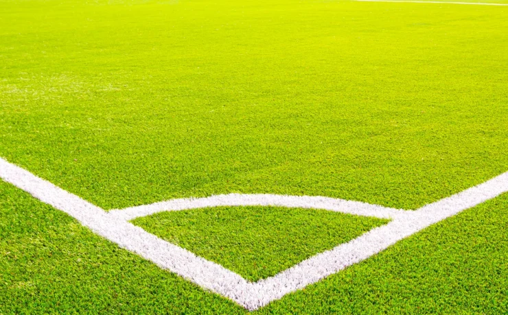 Corner of a green artificial turf soccer field with white lines. Professional football stadium background under bright sunlight. Concept of sports, competition, and outdoor activity.