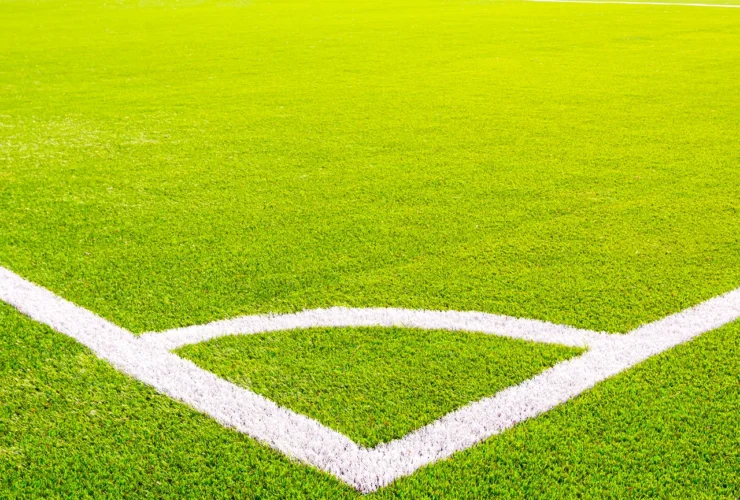 Corner of a green artificial turf soccer field with white lines. Professional football stadium background under bright sunlight. Concept of sports, competition, and outdoor activity.