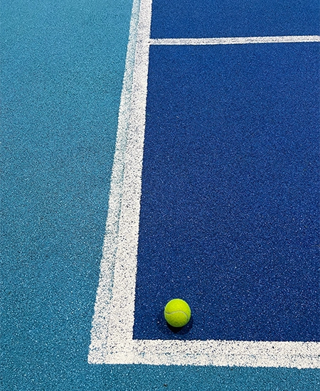 Close-up image of a bright yellow tennis ball placed at the corner of a blue hard court. The crisp white lines and contrasting court colors emphasize precision