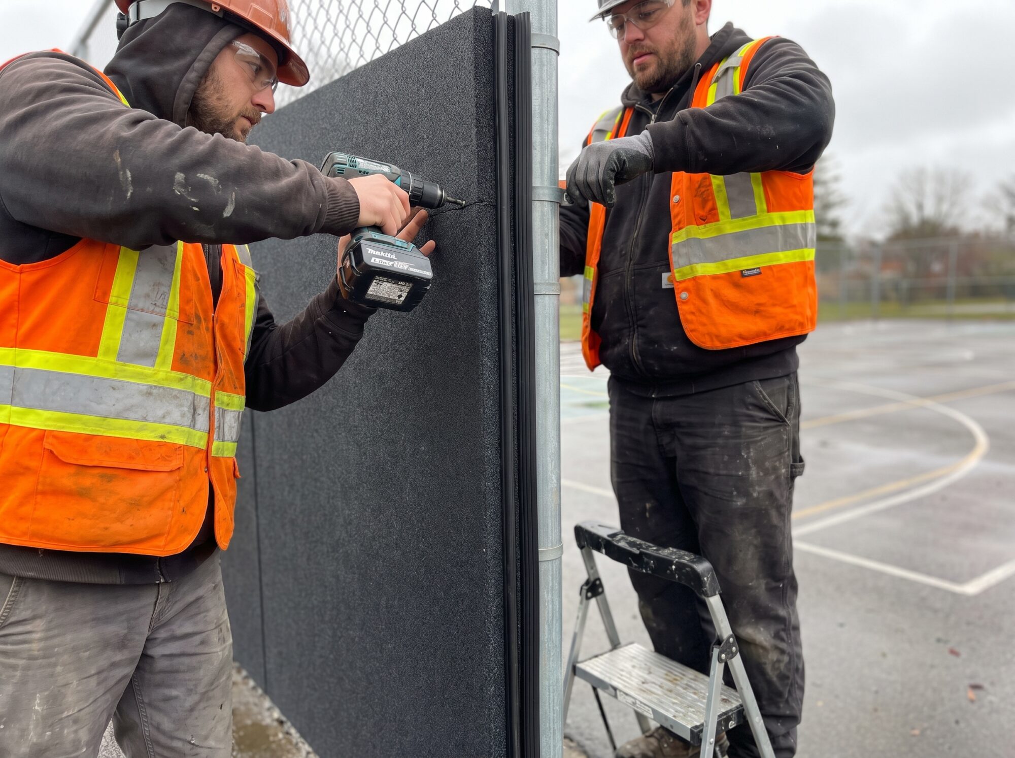 Two contractors in hard hats and hi-vis vests fastening a dark grey acoustic barrier panel to a galvanized fence post beside a sports court