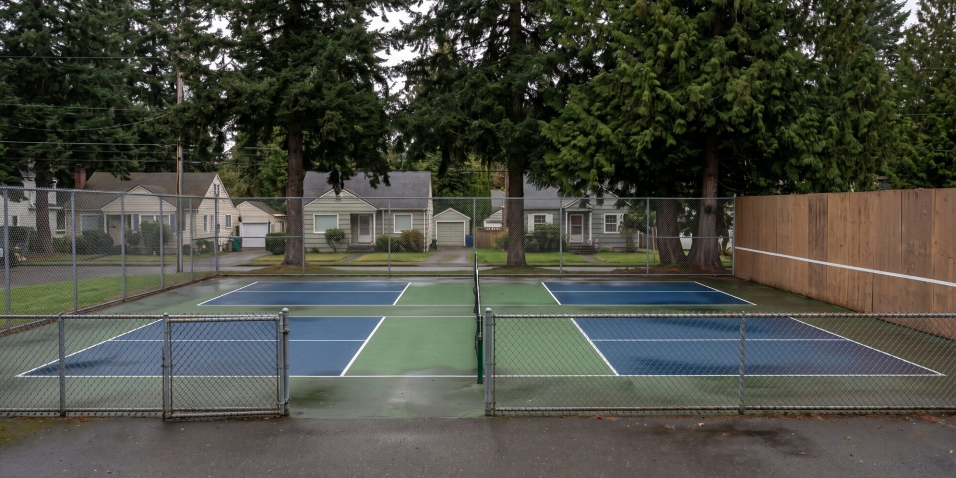 Two outdoor pickleball courts with a solid acoustic barrier panel along the side fence facing nearby houses in an Ontario neighbourhood