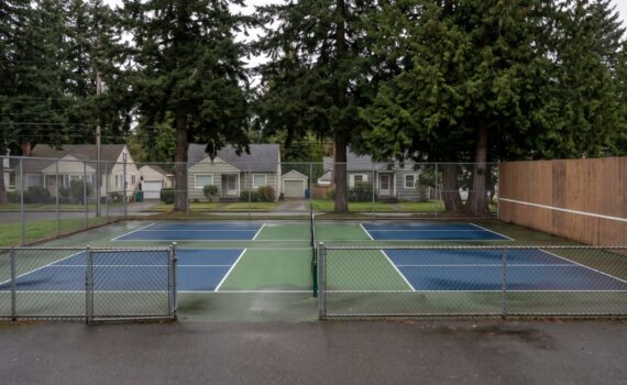 Two outdoor pickleball courts with a solid acoustic barrier panel along the side fence facing nearby houses in an Ontario neighbourhood
