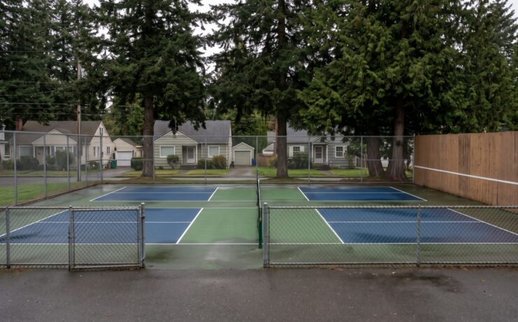 Two outdoor pickleball courts with a solid acoustic barrier panel along the side fence facing nearby houses in an Ontario neighbourhood
