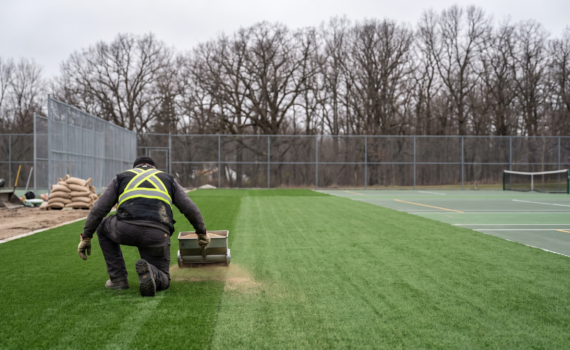 Contractor kneeling on an outdoor artificial turf sports court using a spreader to distribute infill granules