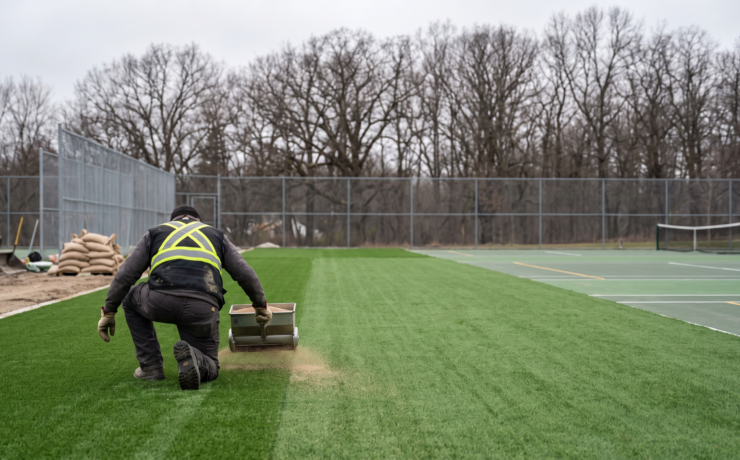 Contractor kneeling on an outdoor artificial turf sports court using a spreader to distribute infill granules