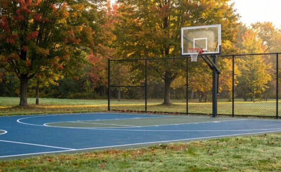 Newly built backyard half-court basketball court with a blue and green acrylic surface, an in-ground hoop pole and black chain-link fence under autumn maples