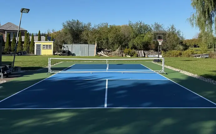 Backyard pickleball court with two-tone blue playing surface, green border, portable net and basketball hoop