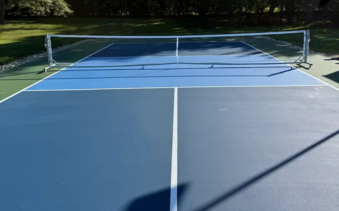 Two-tone blue backyard pickleball court with white lines and portable net, framed by trees