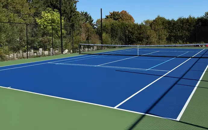 Newly coated blue tennis court with pickleball lines, green surround and black chain-link fence among trees