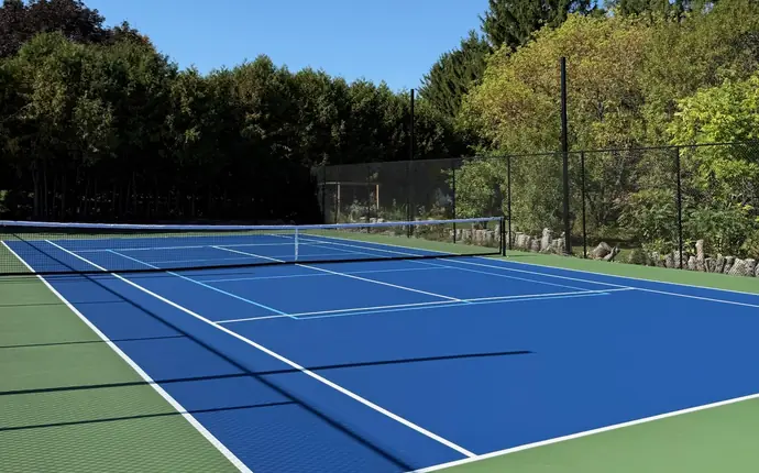 Blue and green tennis court with pickleball lines and fence shadows, bordered by evergreen trees