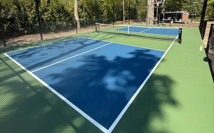 Backyard pickleball court with basketball hoop, black chain-link fence and mature trees