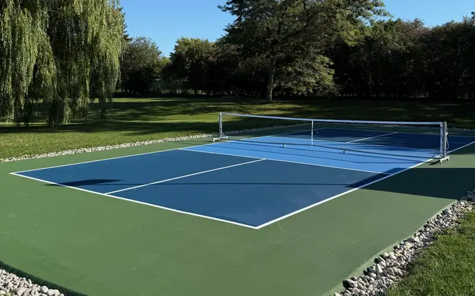 Backyard pickleball court edged with river stone, with a willow tree and open lawn behind it