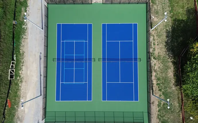 Overhead drone view of two blue tennis courts on a green surround, one with blended pickleball lines